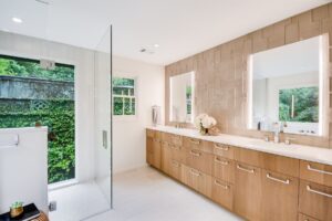 Modern bathroom with double sinks, wood cabinets, large mirrors, and a glass shower next to windows overlooking greenery.