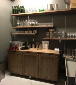 Wooden shelves with glasses, bottles, and bar tools above a cabinet and countertop in a dimly lit room.