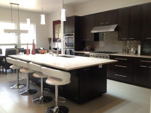 Modern kitchen with dark cabinets, white countertops, bar stools, and stainless steel appliances, brightly lit by windows.