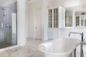 Modern bathroom with a freestanding tub, glass shower, marble floors, and white cabinets with glass doors.