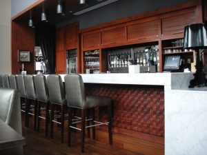 A modern bar with a marble counter, high chairs, hanging lights, and shelves stocked with bottles and glassware.