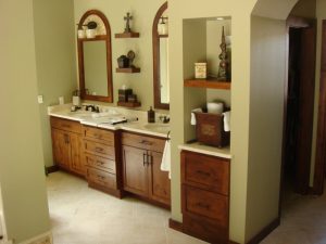 Bathroom with wooden cabinets, double sinks, mirrors, shelves with decor, and a towel holder on beige tiled floor.