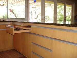 Light wood kitchen cabinets with two drawers open beneath a dark countertop, large windows in the background.