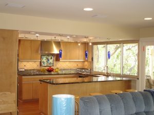 Modern kitchen with wood cabinets, granite countertops, blue pendant lights, and large windows with a view of trees.