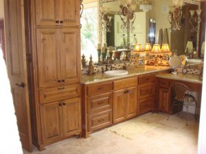 A bathroom with wooden cabinets, two sinks, mirrors, decorative lights, and a cushioned chair on tiled flooring.