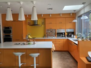 Modern kitchen with bright orange cabinets, white countertops, bar stools, and stainless steel appliances.