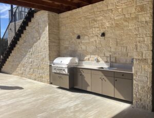 Outdoor kitchen with stone wall, built-in grill, sink, cabinets, and overhead lights under a staircase.