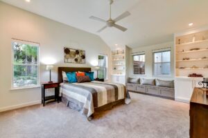 Bright bedroom with a striped bedspread, built-in shelves, window seat, ceiling fan, and carpeted floor.