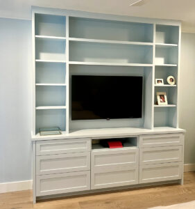 Built-in white entertainment center with open shelves, a flat-screen TV, framed photos, and drawers below on a wooden floor.