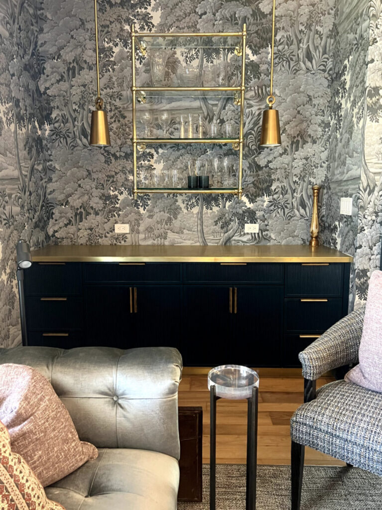 A black sideboard with gold accents stands below glass shelves, flanked by pendant lights and patterned wallpaper.