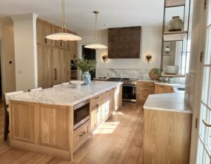 Modern kitchen with light wood cabinets, marble countertops, pendant lights, and stainless steel appliances.
