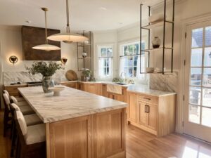 Kitchen with light wood cabinets, marble countertops, open shelves, modern lighting, and large windows for natural light.