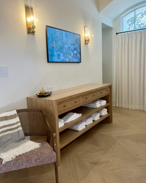 Wooden console with folded towels, a tray, and wall art in a bright room with a chair and curtains.