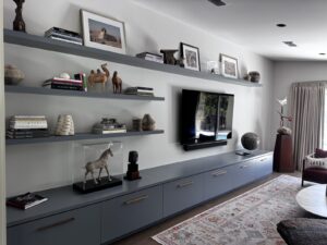 Modern living room with gray shelves, books, art, and sculptures, a wall-mounted TV, and a patterned rug.