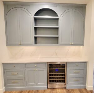 Light blue kitchen cabinets with gold handles, open shelves, marble backsplash, and a wine fridge beneath the counter.