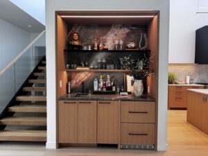 A modern home bar with wooden cabinets, a small sink, shelves of liquor bottles, and decorative items on a marble backsplash.