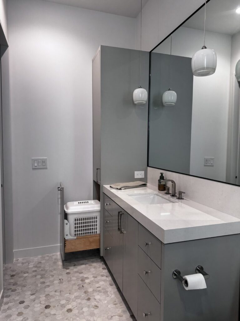 Modern bathroom with gray cabinets, a large mirror, pendant lights, and a laundry basket in a pull-out drawer.