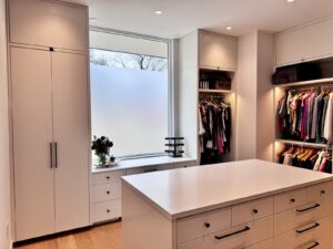 Modern walk-in closet with white cabinets, center island, hanging clothes, and a large frosted window.