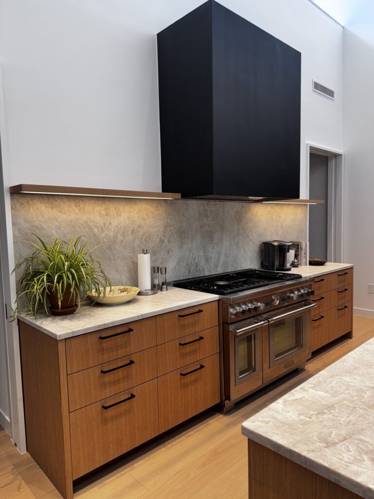 Modern kitchen with wood cabinets, marble countertop, stainless steel stove, black range hood, and a potted plant.
