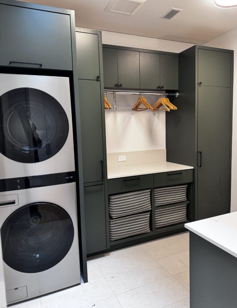 Modern laundry room with stacked washer and dryer, green cabinets, countertop, and yellow hangers on a rail.