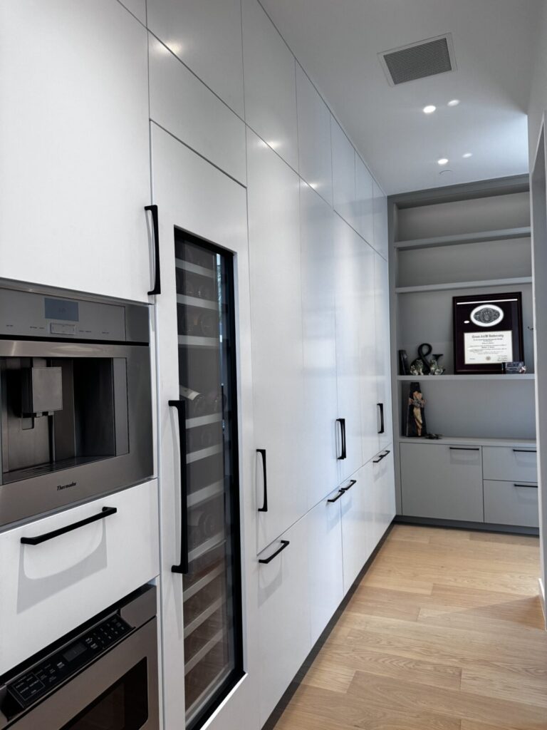 Modern kitchen with white cabinets, built-in appliances, wood floor, and a wine fridge; shelves hold a framed certificate.