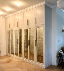 Cream-colored built-in cabinets with mirrored doors and long handles in a well-lit room with wood herringbone flooring.