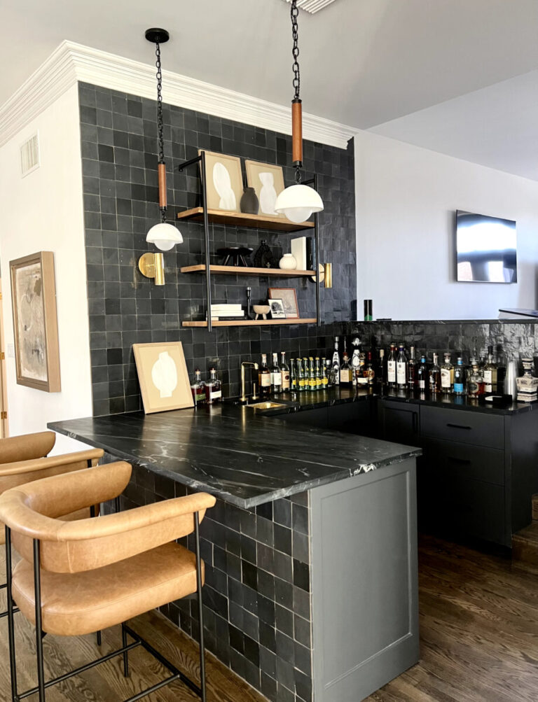 Home bar with black tiled backsplash, shelves with abstract art, liquor bottles, and tan barstools on a wooden floor.