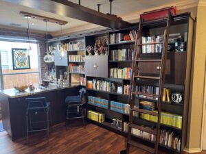 Home library with dark wood shelves, books, collectibles, a ladder, bar area, and two barstools by a window.