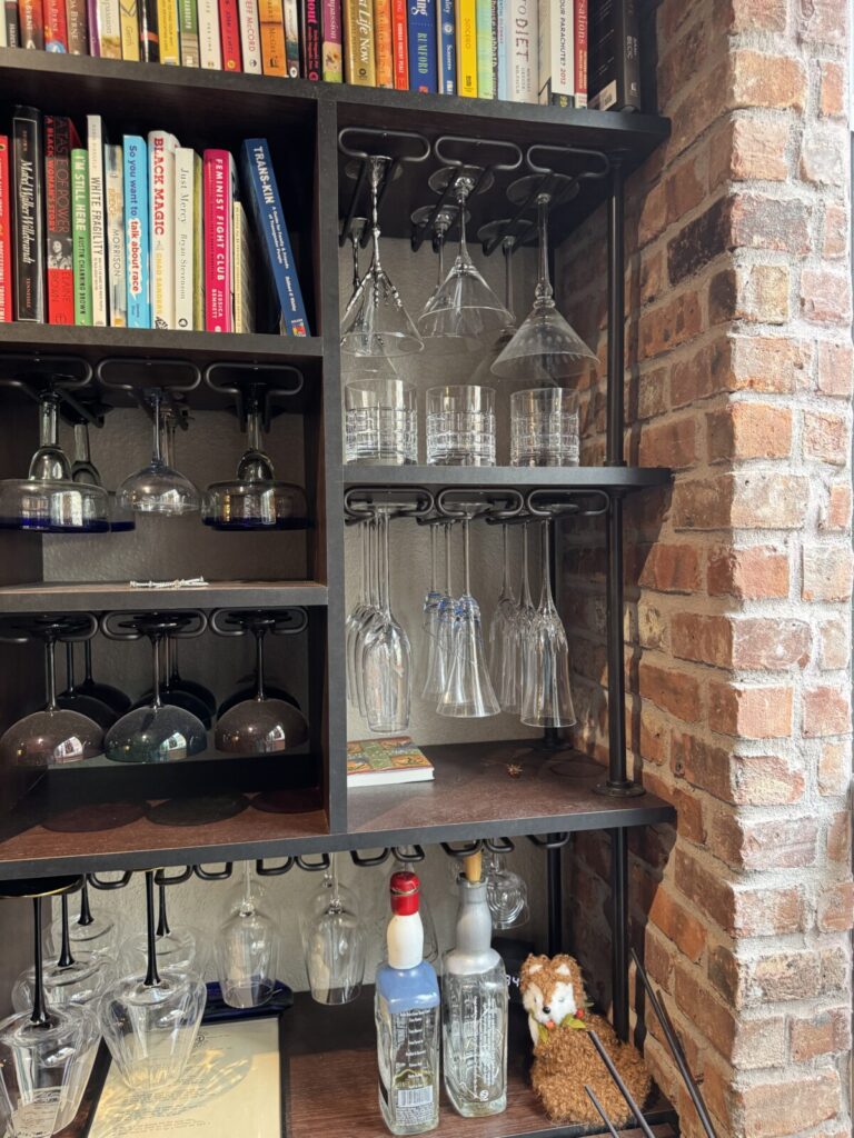 Books and glassware on shelves, with various glasses hanging upside down and bottles on the lower shelf near a brick wall.