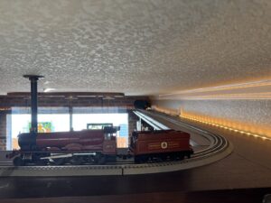 A model red steam train with Hogwarts branding sits on a track beneath a low ceiling with LED strip lights.