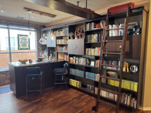 Home library with bookshelves, a ladder, a bar counter, two chairs, and a large window with stained glass.