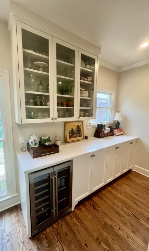 White cabinets with glass doors, a wine fridge below, and decorative items arranged on a white countertop.