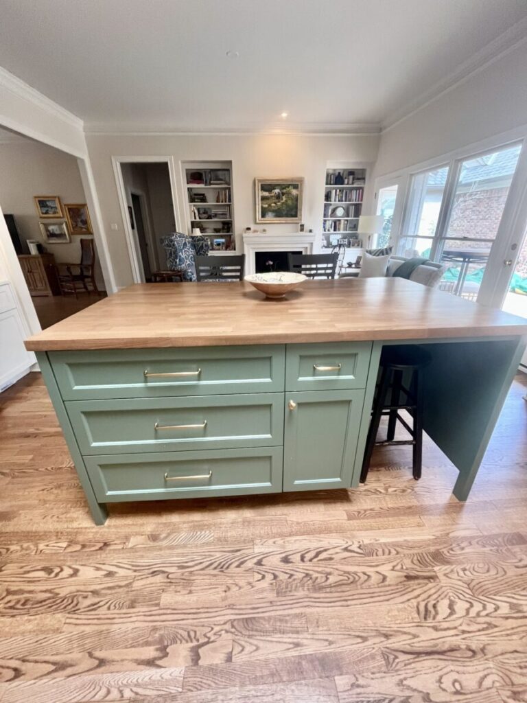 A green kitchen island with gold handles, a wood countertop, and stools stands on a hardwood floor in a bright, open room.