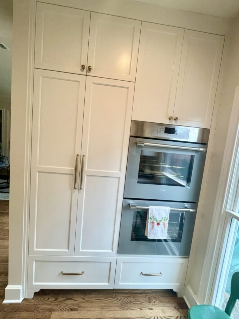 White built-in kitchen cabinets next to stacked stainless steel wall ovens with a towel hanging on the oven handle.