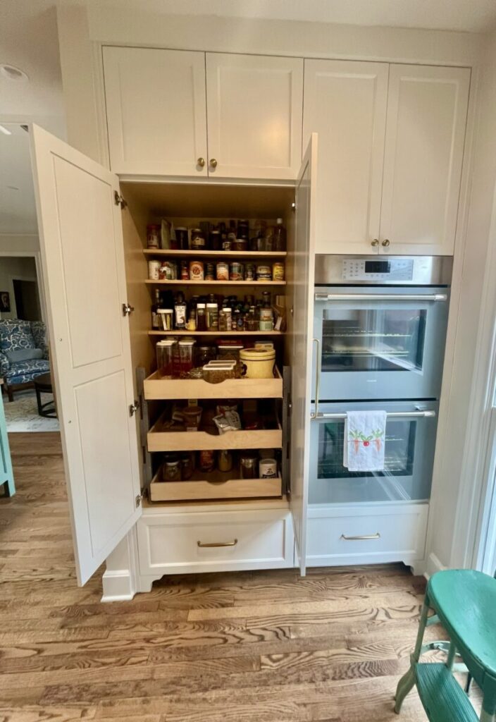 Open pantry cabinet with pull-out shelves holding jars, cans, and food items, next to a double wall oven in a kitchen.