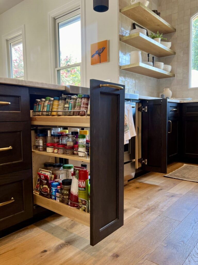 A kitchen with dark cabinets, wooden floors, and a pull-out pantry drawer open, displaying spices and canned goods.