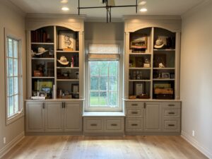 Built-in shelves and cabinets surround a window seat, displaying books, hats, photos, and decorative items.