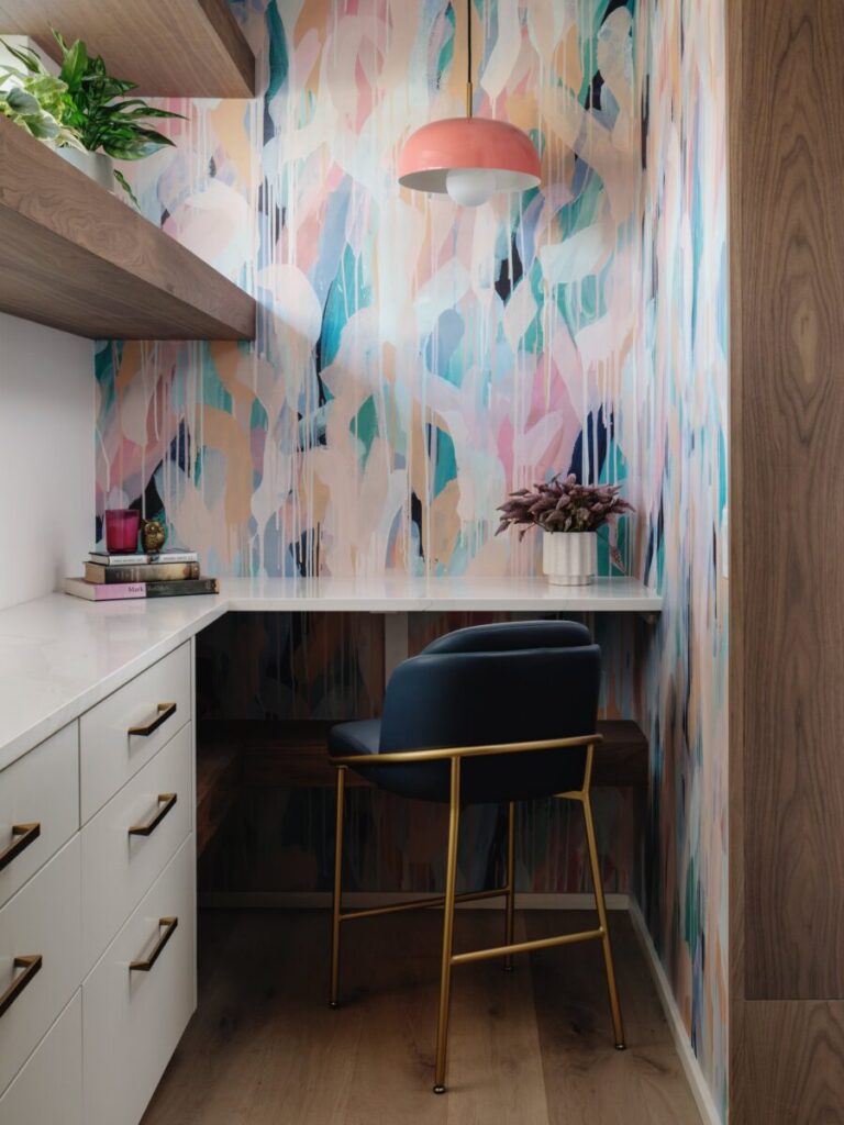 Small home office with abstract pastel wallpaper, a white desk, black chair with gold legs, and wooden shelves.