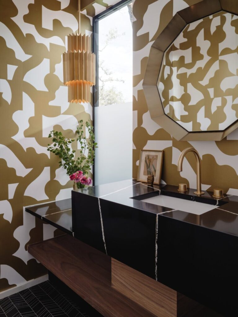 Bathroom with bold abstract wallpaper, black sink, gold fixtures, geometric mirror, and a vase with flowers on the counter.