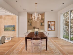 Modern dining room with a dark wood table, clear chairs, chandelier, wall art, shelves, and large window.
