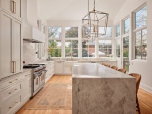 Bright kitchen with marble island, white cabinets, large windows, stainless appliances, and woven chairs along the counter.