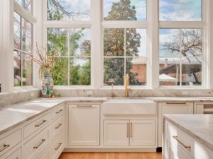 Bright kitchen with white cabinets, marble countertops, gold hardware, a farmhouse sink, and large windows with garden views.
