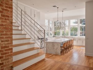 Modern kitchen with marble island, wooden floors, large windows, white cabinets, and adjacent staircase with metal railing.