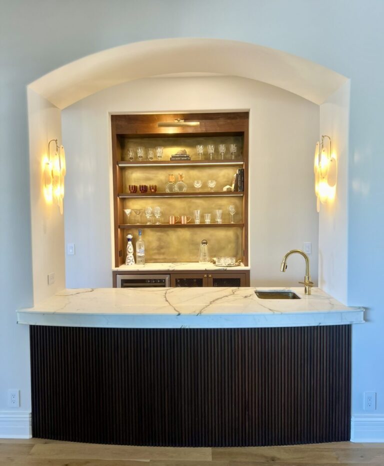 Home bar with marble countertop, gold faucet, glass shelves with glassware, and wall sconces under an arched alcove.