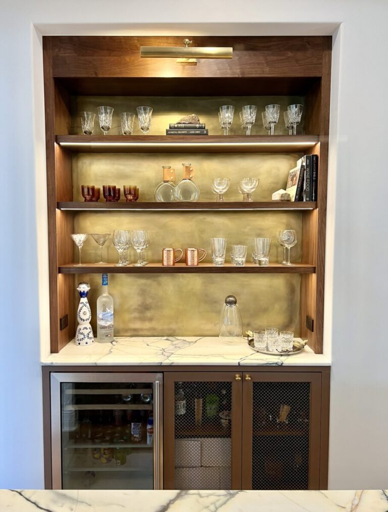 Built-in bar with shelves holding assorted glassware, bottles, and mugs above a marble countertop and beverage fridge.