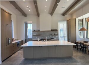 Modern kitchen with a large island, wood cabinetry, exposed ceiling beams, and ample natural light from tall windows.