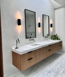 Modern bathroom with dual sinks, wood vanity, two mirrors, wall lights, and a small potted plant on marble flooring.