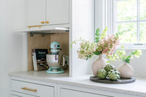 A kitchen counter with a mixer, cookbooks, vases of flowers, and artichokes beside a window.