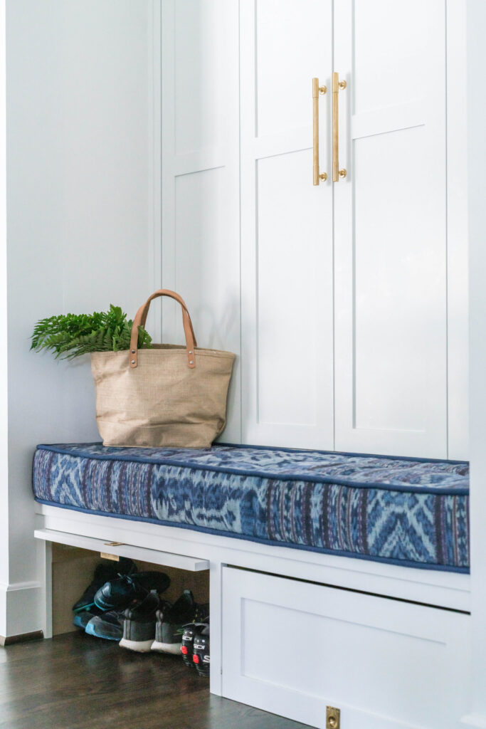 Blue cushioned bench with a beige tote bag, green plant, and storage space holding shoes underneath.
