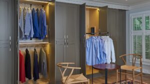 Open closets show neatly hung shirts and jackets, with two chairs and a small round table in front.